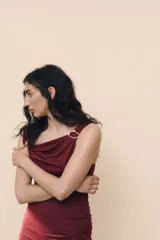 Vestido midi color vino tinto, sin mangas, con escote drapeado tipo cascada y un detalle de argolla metálica plateada en el bretel derecho. Presenta frunces laterales a lo largo del torso.