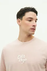 Remera verde de manga corta con cuello redondo y estampado en la espalda con la frase "Côte d'Azur Surfer avec les mouettes" y un dibujo de una sombrilla, una reposera y gaviotas.