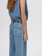 Mono largo de jean celeste con corte holgado, cintura elastizada con cordón ajustable, sin mangas, cuello camisero y cierre frontal con botones. Presenta bordados de motivos playeros en el pecho.