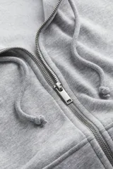 Campera con capucha gris melange de felpa con interior cepillado suave, hombros caídos, capucha forrada en punto con cordón para ajustar, cierre frontal y bolsillo canguro. Puños y bajo en punto elástico acanalado.