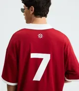 Remera de manga corta estilo polo, color rojo, con cuello y puños en contraste blanco. Presenta un corte oversized con hombros caídos y la palabra "ATHLÈTES" estampada en blanco en el frente. Incluye pequeños gráficos blancos a cada lado del pecho.