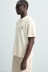 Camiseta color crudo de corte relaxed fit, con cuello redondo y mangas cortas. Presenta un pequeño bordado de flores en el pecho y un estampado fotográfico en la espalda.