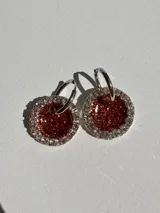 Par de aros colgantes con argolla de acero quirúrgico y dije circular de resina color terracota con glitter, bordeado con strass plateado.