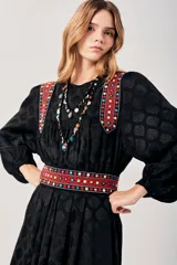 Vestido largo negro con estampado de hojas en relieve, mangas largas abullonadas y falda plisada. Presenta detalles bordados en rojo, blanco y azul en los hombros y la cintura.