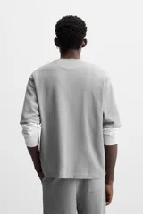 Camiseta gris de corte relaxed fit, cuello redondo y manga corta. Tiene un parche rectangular con texto en el pecho.