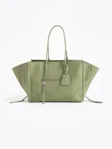 Bolso tote pequeño de cuero vacuno color verde claro, con forma trapezoidal y asas de mano. Presenta cierres decorativos y funcionales en los laterales que permiten expandir el bolso, un bolsillo frontal con cierre y un tag colgante con aplique metálico.