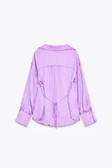 Camisa lila de satén con cuello solapa, escote en pico y manga larga. Tiene detalle de lazada en la espalda, bajo con aberturas laterales y cierre frontal con botones.
