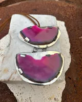 Collar con colgante de dos piedras naturales irregulares engarzadas en metal plateado y dorado, suspendidas de un cordón de cuero marrón con detalle de nudo corredizo. Las piedras tienen vetas en tonos naranja, marrón y blanco.