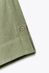 Camisa verde oliva confeccionada con lino, de corte cropped, con cuello solapa, escote pico, manga corta con trabilla y dos bolsillos delanteros de plastrón. Cierre frontal con botones.