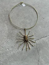 Gargantilla tipo collar rígido de metal dorado, con un dije colgante que simula un sol o estrella con un cristal rectangular central de tonos ámbar y negro.