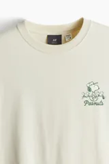 Remera color crema de corte holgado, con cuello redondo y estampado pequeño de Snoopy y la frase "Peanuts" en el pecho.