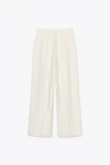 Pantalón fluido color crudo, de tiro medio con cintura con trabillas, bolsillos laterales, pinzas y raya planchada en delantero. Cierre frontal con cremallera, botón y ganchos metálicos.