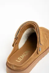 Zueco tipo mule de gamuza color camel, con forro interior de corderito visible en el borde. Presenta una tira ajustable con broche metálico en el lateral y suela de goma gruesa y dentada.