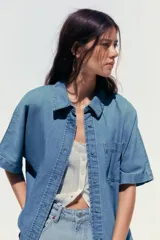 Camisa de denim celeste con cuello solapa, manga corta, bolsillo de parche en el pecho, bajo con aberturas laterales y cierre frontal con botones.