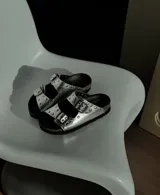 Sandalia plateada de cuero vacuno con forrería vacuna y detalle de tachas en la capellada. Tiene una plantilla birk forrada en cuero, y fondo negro de eva.