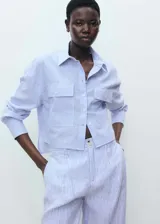 Camisa corta a rayas verticales celestes y blancas, de manga larga con puños abotonados, cuello camisero, cierre frontal con botones, y dos bolsillos de parche con solapa en el pecho.