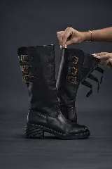 Bota biker negra de caña media, confeccionada en cuero liso, con suela gruesa y taco bajo estriado. Presenta tres hebillas doradas decorativas en el lateral de la caña y tiras ajustables en la parte superior.