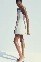 Vestido corto blanco de tejido elástico, con escote recto, tirantes finos ajustables, detalle de pliegues y cierre con cremallera oculta en la costura.