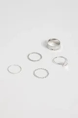 Set de cinco anillos plateados de diferentes diseños: uno ancho con textura martillada, uno liso, uno con pequeñas esferas, uno con forma de triángulo y uno con diseño trenzado.