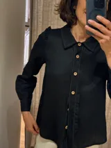 Camisa negra de lino con corte femenino, cuello camisero, manga larga abullonada con puño y volados en los hombros. Cierre frontal con botones de madera.
