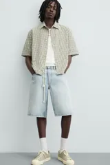 Bermuda de denim de algodón, color gris claro lavado, de corte ancho (baggy fit) y largo hasta la rodilla. Presenta cinco bolsillos y cierre frontal con cremallera y botón.