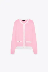 Chaqueta de punto rosa con cuello redondo y manga larga. Tiene falsos bolsillos delanteros y cierre frontal con botones dorados. Los bordes del cuello, los bolsillos y el bajo son blancos.