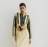 Camisa blanca con rayas verticales negras, de manga larga y corte recto. Tiene cuello clásico, cierre con botones y bolsillo en el pecho.