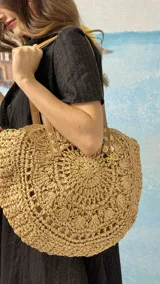 Bolso playero grande de tejido de papel trenzado color paja, con forma redonda y asas de cuero marrón. Presenta un forro interior y un dije colgante con motivos marinos.