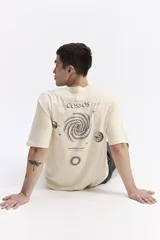 Remera oversize de manga corta color beige, con escote redondo y estampado trasero de temática espacial que incluye la palabra "COSMOS", una galaxia en espiral, planetas y estrellas.