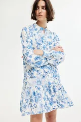 Vestido corto de viscosa con estampado floral en tonos azules. Tiene cuello mao, botones frontales, mangas largas abullonadas con puños abotonados y falda con volantes.