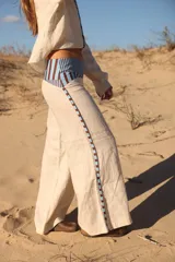 Pantalón de lino color crudo, de corte ancho y tiro medio. Presenta detalles de rayas verticales en color marrón y celeste en los laterales y en la cintura.