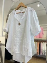 Blusa color crema con cuello camisero, mangas cortas con dobladillo y cierre frontal con un botón de madera. Presenta bordados con motivos de paisley y perforaciones.