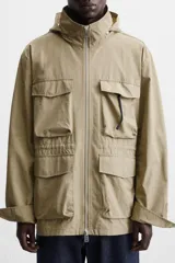 Parka color beige de tejido técnico ligero, corte relaxed fit, cuello alto con capucha plegable, manga larga con puños ajustables con botón a presión, cierre frontal con cremallera, cintura ajustable con elástico interno y múltiples bolsillos frontales de parche con solapa.