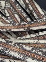 Cinturón de cuero con estampado de piel de serpiente en tonos blanco, gris y negro. Presenta una hebilla ovalada de metal color bronce y detalles de costuras en hilo color cobre.
