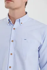 Camisa celeste de tejido texturado oxford, corte clásico, con cuello abotonado, mangas largas con puños ajustables, bolsillo en el pecho y cierre frontal con botones de madera.