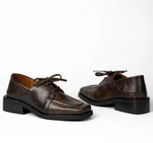 Zapato acordonado de cuero vacuno color marrón oscuro, con suela de goma, horma cuadrada y tacón bajo.