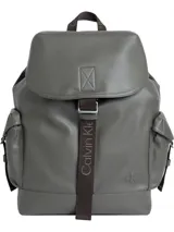 Mochila Calvin Klein de poliuretano en color gris con textura granulada. Presenta un diseño funcional con solapa superior y cierre mediante una correa de tela marrón con el nombre de la marca. Incluye bolsillos laterales tipo cargo con solapa, asa superior y el logo 'ck' grabado en relieve en la esquina inferior frontal.