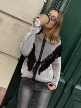 Campera tejida con textura vanizada a rayas horizontales en blanco y negro, con cierre frontal y flecos negros en forma de V en el pecho.