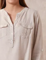 Blusa color beige de lino y rayón, con cuello a la base, escote en V y abertura con botones. Tiene bolsillos en la parte delantera y mangas largas regulables con presilla y botón.