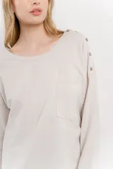 Remera color beige de corte holgado, con mangas largas, cuello redondo y bolsillo en el pecho. Presenta detalle de botones en el hombro.