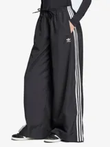 Pantalón deportivo negro de corte ancho con tres rayas blancas verticales en los laterales y logo de Adidas bordado en blanco en la parte superior de la pierna izquierda.