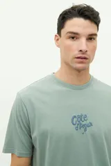 Remera verde de manga corta con cuello redondo y estampado en la espalda con la frase "Côte d'Azur Surfer avec les mouettes" y un dibujo de una sombrilla, una reposera y gaviotas.