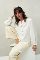Camisa blanca de manga larga con botones frontales y cuello mao. Presenta pequeños pliegues en los hombros y detalle calado sutil.