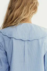 Camisa celeste a rayas verticales blancas, de corte recto, con cuello bobo con bordados y escote pico con lazada. Tiene mangas cortas abullonadas con elástico y cierre frontal con botones.