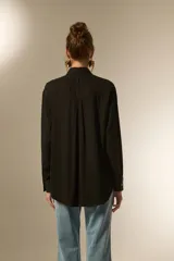 Camisa holgada de manga larga, color negro, con cuello camisa y botones frontales. Presenta apliques de tachas plateadas en los hombros y a lo largo de la tapeta frontal.