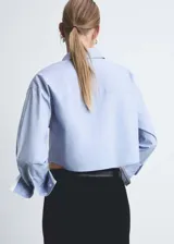 Camisa celeste de popelina de algodón con diseño crop, cuello camisero, cierre frontal con botones, dos bolsillos de solapa en la parte delantera, manga larga con puños abotonados y terminaciones en contraste blanco.