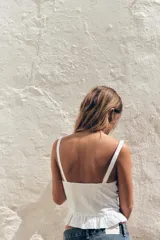 Top blanco estilo corsé con escote corazón, tirantes finos y bordados a tono. Presenta un bajo con volantes y cierre frontal con ganchos metálicos. La espalda es de tejido elástico tipo nido de abeja.