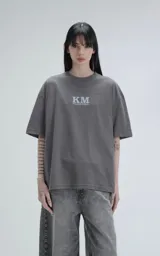 Remera de algodón premium color gris concreto, con corte oversize, hombros caídos y cuello redondo. Presenta un estampado frontal con las siglas "KM" y el texto "#KILOMBITOMAL" debajo en color celeste pálido.