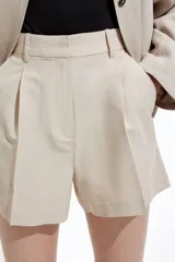 Short color beige de tiro alto con pliegues delanteros, bolsillos al bies y cierre frontal con botón y corchete ocultos.