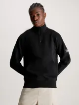Buzo negro de felpa de algodón orgánico con cuello alto con cremallera, puños y bajo de canalé, bolsillo canguro y parche con logo en la manga.